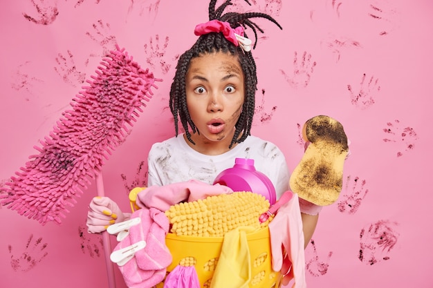 Geschokt jong etnisch dienstmeisje kijkt naar een erg vuile kamer met chaos rond houdt spons en dweil bezig met opruimen keuken verzamelt wasgoed in mand doet huisklusjes poseert tegen roze muur