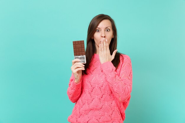 Geschokt jong meisje in gebreide roze trui in de hand houden chocoladereep die mond bedekt met palm geïsoleerd op blauwe turquoise muur achtergrond studio portret. Mensen levensstijl concept. Bespotten kopie ruimte.