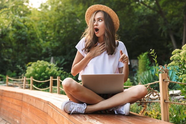 Geschokt jong meisje zit met laptopcomputer in het park buitenshuis, wijzende vinger