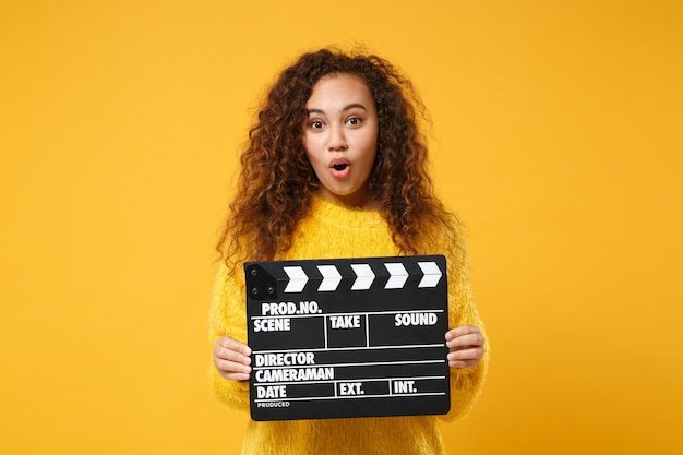 Geschokt jonge Afro-Amerikaanse meisje in bont trui poseren geïsoleerd op geel oranje achtergrond in studio. Mensen levensstijl concept. Bespotten kopie ruimte. Klassieke zwarte film filmklapper vasthouden.