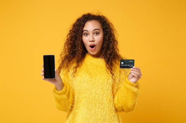Geschokt jonge Afro-Amerikaanse meisje in bont trui poseren geïsoleerd op gele achtergrond. Mensen levensstijl concept. Bespotten kopie ruimte. Mobiele telefoon vasthouden met leeg leeg scherm, creditcard.