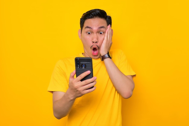 Geschokt jonge Aziatische man in casual tshirt kijken naar mobiele telefoon scherm reageren op online nieuws geïsoleerd op gele achtergrond Mensen lifestyle concept