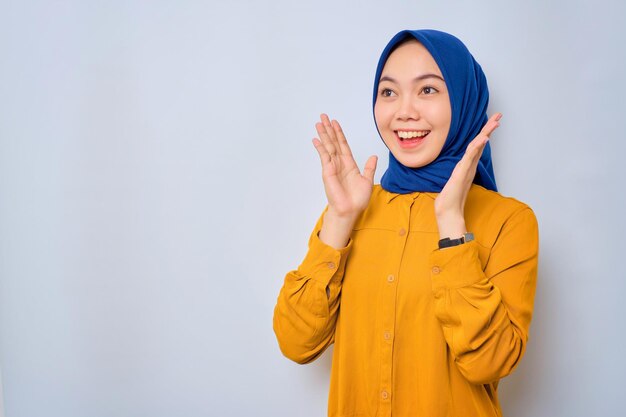 Geschokt jonge Aziatische moslimvrouw gekleed in oranje shirt kijken naar kopie ruimte en het verhogen van handen met open mond reageren op verkoop aanbod geïsoleerd op witte achtergrond