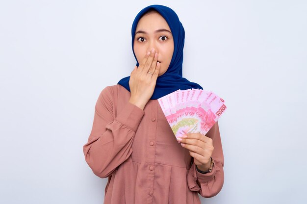 Geschokt jonge Aziatische moslimvrouw in roze shirt met geld bankbiljetten geïsoleerd op witte achtergrond Mensen religieuze levensstijl concept