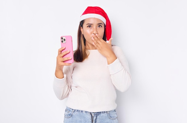 Geschokt jonge Aziatische vrouw in een kerstmuts met mobiele telefoon die mond bedekt met de hand geïsoleerd op een witte achtergrond