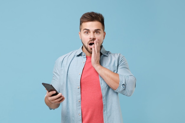 Geschokt jonge, bebaarde man in casual shirt poseren geïsoleerd op pastel blauwe muur achtergrond studio portret. Mensen emoties levensstijl concept. Bespotten kopie ruimte. Gebruik de mobiele telefoon en leg de hand op de wang.