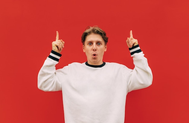 Geschokt jonge knappe man in wit sweatshirt staat op rode muur achtergrond en toont duimen omhoog
