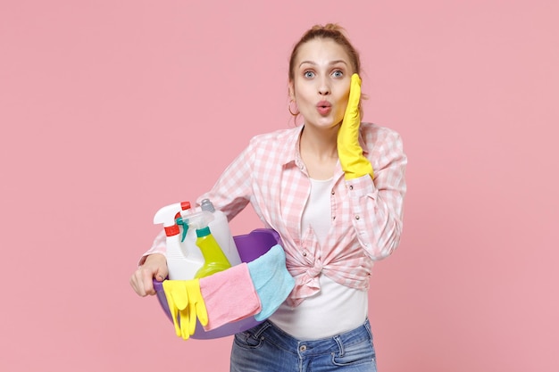 Geschokt jonge vrouw huisvrouw in rubberen handschoenen houden bekken met wasmiddelflessen wassen reinigingsmiddelen terwijl het doen van huishoudelijk werk geïsoleerd op roze achtergrond studio. Huishoudelijk concept. Leg de hand op de wang.