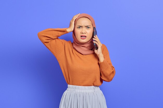 Geschokt mooie Aziatische vrouw in bruine trui praten op mobiele telefoon met open mond geïsoleerd over paarse achtergrond. Mensen islam religieus concept