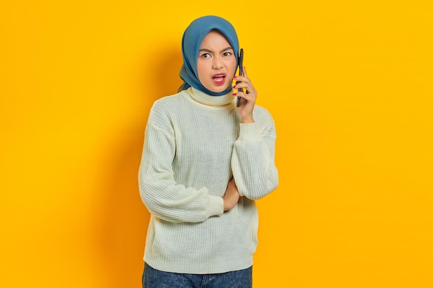 Geschokt mooie Aziatische vrouw in witte trui en hijab praten op mobiele telefoon terwijl ze opzij kijkt geïsoleerd over gele achtergrond