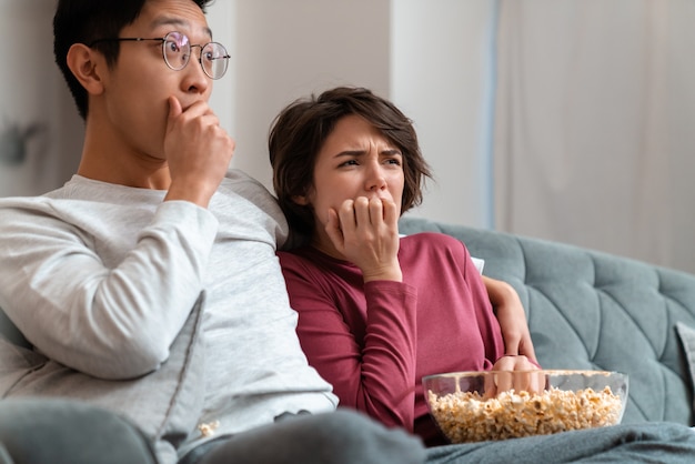 Geschokt multinationaal stel dat popcorn eet en film kijkt terwijl ze thuis op de bank zitten