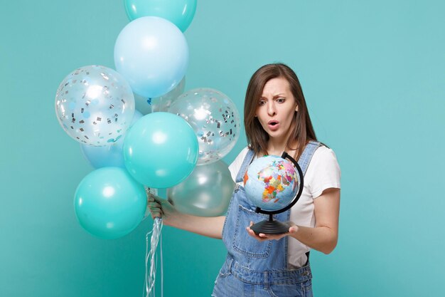 Geschokt perplex jonge vrouw kijken op aarde wereldbol in de hand, vieren, houden kleurrijke luchtballonnen geïsoleerd op blauwe turquoise muur achtergrond. Vakantie verjaardagsfeestje, mensen emoties concept.