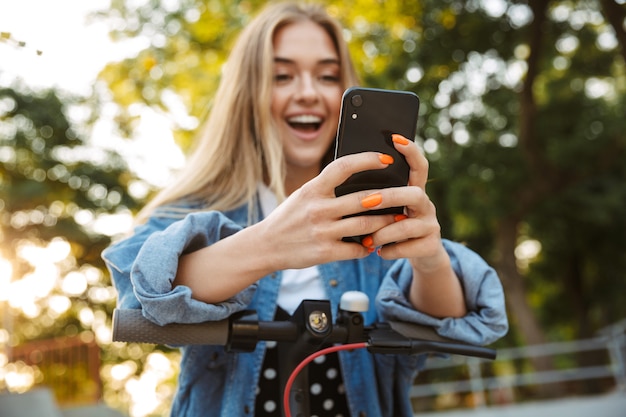 geschokt positief emotioneel tienermeisje in park lopen op scooter met behulp van mobiele telefoon. Focus op telefoon.