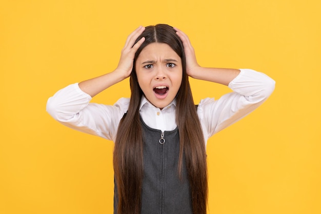 Geschokt tienermeisje in schooluniform neemt hoofd in handen gele achtergrond schok