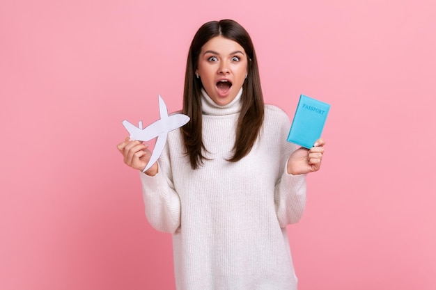 Geschokt verbaasde jonge vrouw met paspoort en papieren vliegtuigje in handen die vakantie in het buitenland plant, gekleed in een witte casual stijl trui Indoor studio opname geïsoleerd op roze achtergrond