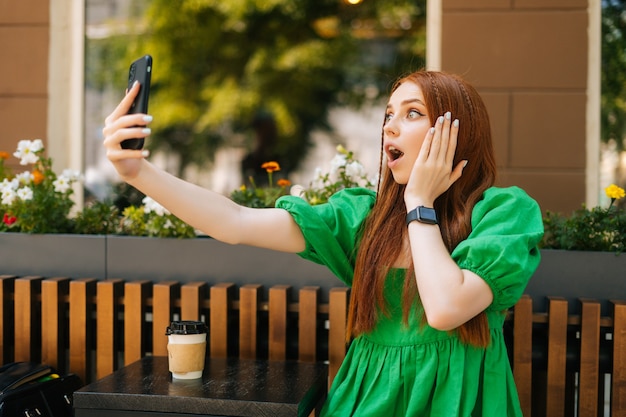 Geschokt verbaasde jonge vrouw met video online chat via camera mobiele telefoon zittend aan tafel