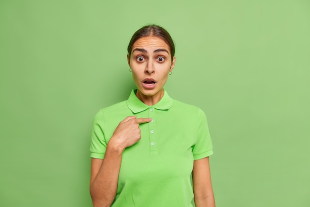 Geschokt verontwaardigde jonge vrouw wijst naar zichzelf met afgeluisterde ogen en geopende mond kijkt met ongeloof gekleed in casual t-shirt geïsoleerd over felgroene muur