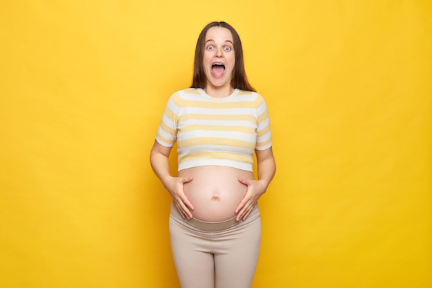 Geschokt verrast blanke zwangere vrouw met blote buik die casual top draagt geïsoleerd over gele achtergrond haar buik vasthoudt schreeuwend begin van samentrekkingen