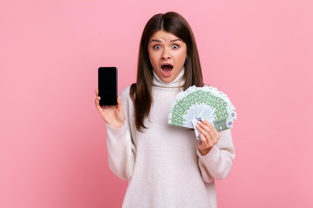 Geschokt verraste vrouw met donker haar met eurobankbiljetten en smartphone met een leeg scherm, met een witte casual stijltrui. Indoor studio opname geïsoleerd op roze achtergrond.
