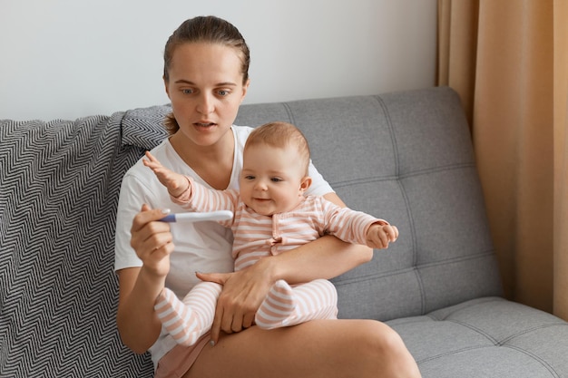 Geschokt verraste vrouw met een staartje met een wit t-shirt dat met haar dochtertje op de bank zit en een zwangerschapstest in handen houdt, verbaasd over het positieve resultaat.