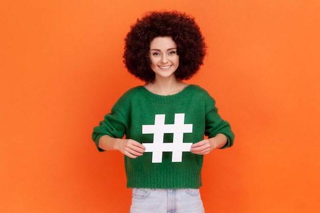 Foto geschokt vrouw met afro kapsel wijzend op witte hashtag symbool verbazingwekkend bevordering van virale onderwerp in sociaal netwerk tagging blog trends indoor studio shot geïsoleerd op oranje achtergrond