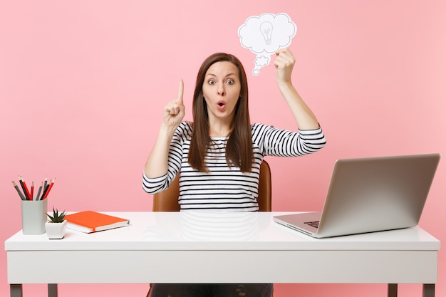 Geschokt vrouw wijzende vinger omhoog houden Say cloud tekstballon met gloeilamp zitten werk op wit bureau met pc-laptop geïsoleerd op pastel roze achtergrond. Prestatie zakelijke carrière concept. Ruimte kopiëren.