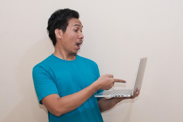 Geschokte Aziatische man met groen T-shirt, casual kleding, laptop vasthoudend en naar laptop wijzend.