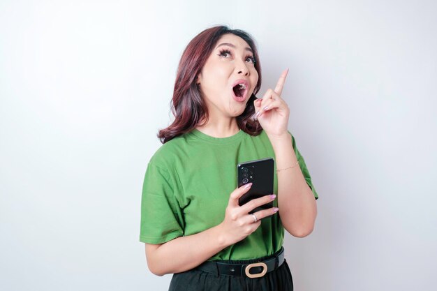 Geschokte Aziatische vrouw die een groen t-shirt draagt dat naar de kopieerruimte wijst en haar telefoon vasthoudt, geïsoleerd door een witte achtergrond