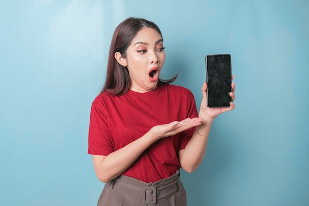 Geschokte Aziatische vrouw die een rood t-shirt draagt dat kopieerruimte op haar smartphone toont en wijst, geïsoleerd door een blauwe achtergrond