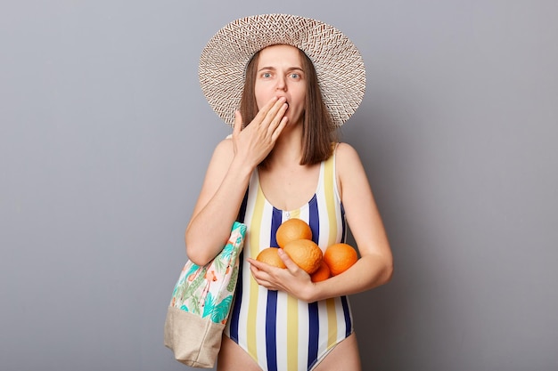 Geschokte, bange vrouw met gestreept zwempak en strohoed die naar de camera kijkt met grote ogen die de mond bedekken met handen geïsoleerde grijze achtergrond met sinaasappels