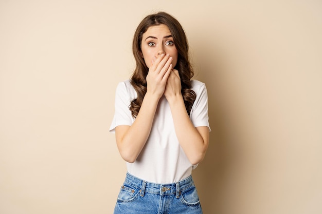 Geschokte brunette vrouw hijgend, sprakeloos naar de camera kijkend, mond bedekken met handen, staande over beige achtergrond