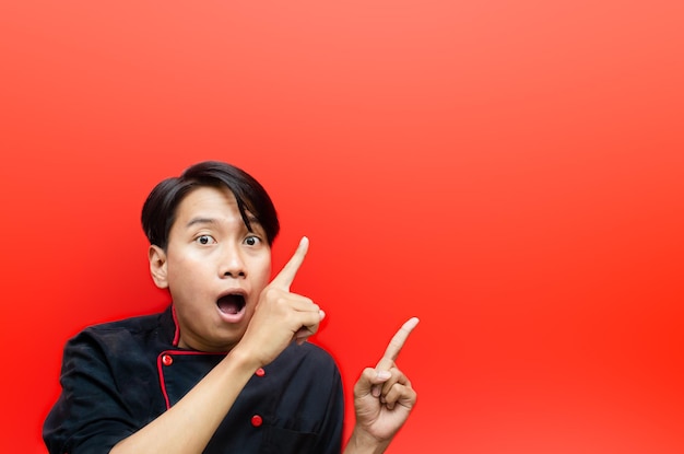 geschokte gelukkig jonge Aziatische chef-kok in zwarte chef-kok uniform iets presenteren over rode achtergrond.