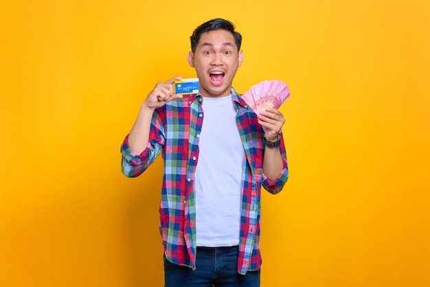 Geschokte jonge Aziatische man in geruit hemd met bankbiljetten en creditcard geïsoleerd op gele achtergrond