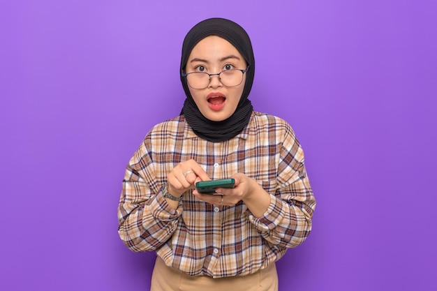 Geschokte jonge Aziatische vrouw in geruit hemd met een mobiele telefoon die geweldig nieuws krijgt en naar camera kijkt geïsoleerd op paarse achtergrond