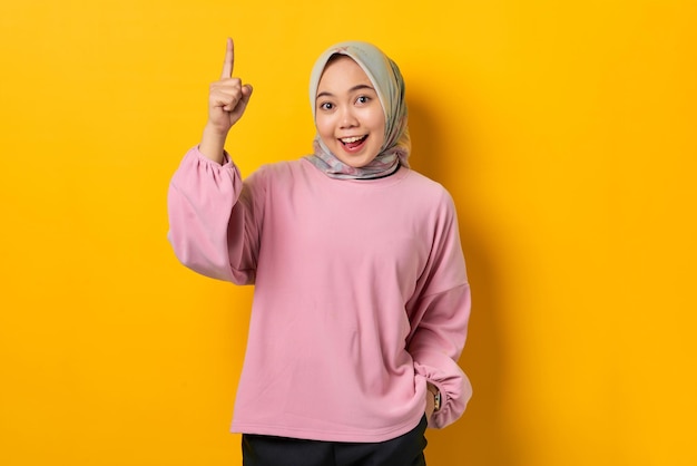 Geschokte jonge Aziatische vrouw in roze shirt die met de vingers omhoog wijst en een goed idee heeft op gele achtergrond