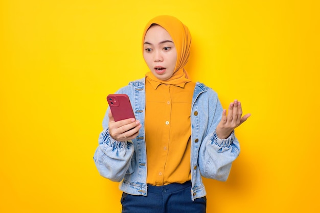 Geschokte jonge Aziatische vrouw in spijkerjasje die naar het scherm van de mobiele telefoon kijkt met open mond die reageert op geweldig nieuws geïsoleerd over gele achtergrond
