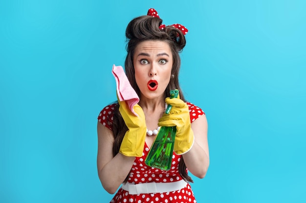 Geschokte jonge pinup-vrouw in retro-outfit met vod en spuitwasmiddel die huishoudelijk werk doet