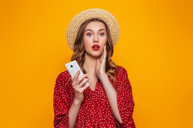 Geschokte jonge vrouw in de rode hoed van het kledingsstro met rode lippenstift die zich met smartphone bevinden en camera bekijken die op een oranje muur wordt geïsoleerd. Verrast meisje met een mobiele telefoon in zomer kleding