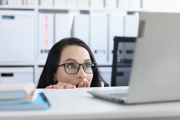 Geschokte jonge vrouw kijkt van onder tafel naar laptopmonitor. Bange vrouw in paniek van internetnieuwsconcept