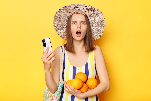 Geschokte verbaasde verbaasde vrouw in gestreept zwempak uit één stuk en strohoed poseren geïsoleerd op gele achtergrond met mobiele telefoon kijkend naar de camera met open mond