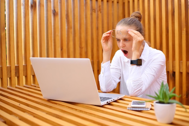 Geschokte verraste vrouw met een wit overhemd zittend in een buitencafé aan het werk op een laptop kijkend naar een notebookmonitor met open mond en grote ogen fout in project