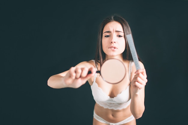 Geschokte vrouw die door een vergrootglas kijkt. Jonge vrouw met lange zwarte geïsoleerde haartribunes. Het meisje heeft een sportief figuur, ze is gekleed in wit ondergoed.