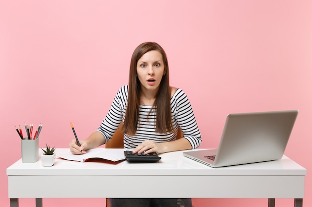 Geschokte vrouw die rekenmachine gebruikt die notities schrijft met berekeningen, zit en werkt op kantoor met een moderne pc-laptop