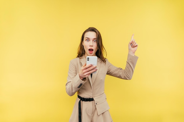 Geschokte vrouw in een beige pak staat met een smartphone in zijn hand op een gele achtergrond