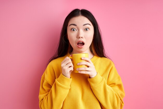 Geschokte vrouw met beker in handen, staat met wijd geopende mond, verrast door iets. geïsoleerd op roze achtergrond