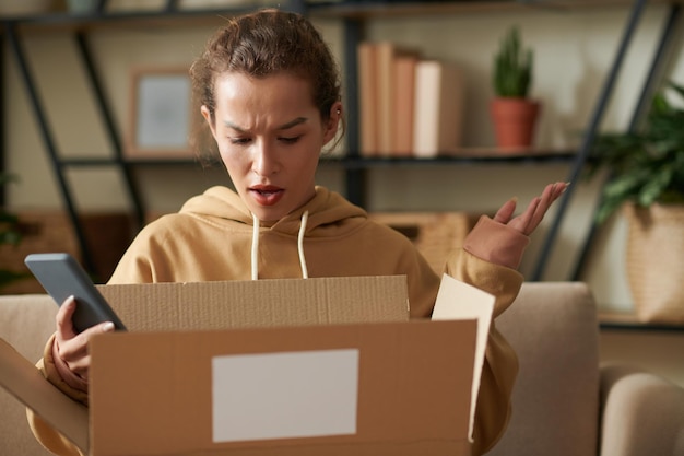 Foto geschokte vrouw met mobiele telefoon en het openen van de kartonnen doos die ze niet leuk vond met de levering van haar pakket