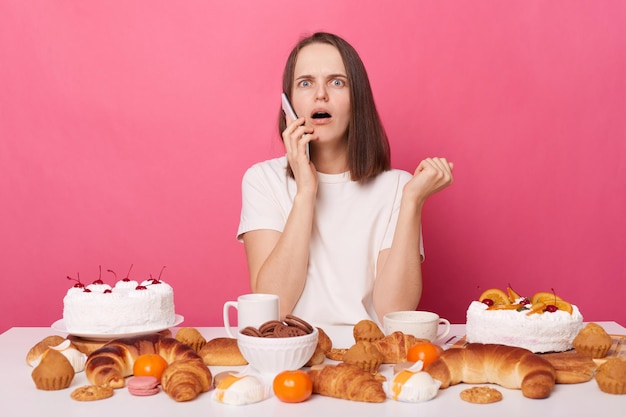 Geschokte vrouw met wit T-shirt zittend aan feestelijke tafel met verschillende desserts geïsoleerd over roze achtergrond pratende telefoon met bange uitdrukking