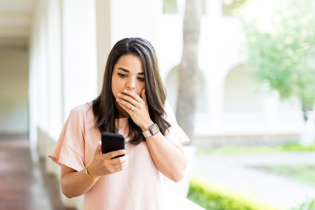 Geschokte vrouwelijke blogger die schokkende informatie leest van internetbronnen op mobiele telefoon in gang