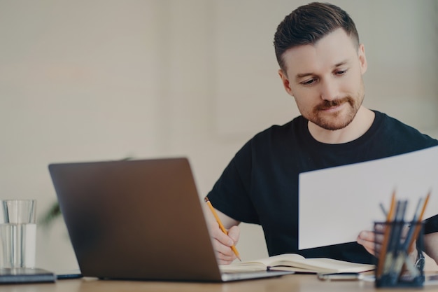 Geschoolde professionele mannelijke expert bezig met het doen van papieren rapportbewerkingen online bestanden via laptop werkt op desktop gericht op documenten gekleed in een casual zwart t-shirt. Bebaarde freelancer op kantoor aan huis