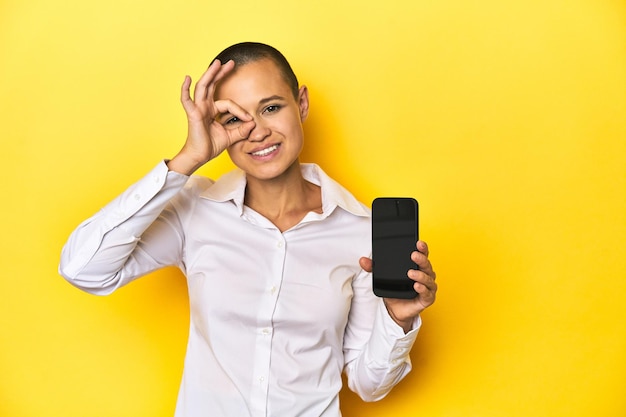 Foto geschoren hoofdvrouw die mobiele gele achtergrond houdt, opgewonden en ok gebaar op het oog houdt